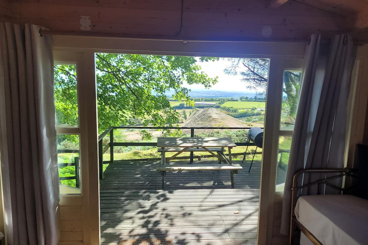 Una stanza con soffitto in legno, un letto, una finestra con tende e vista su un’area picnic esterna.