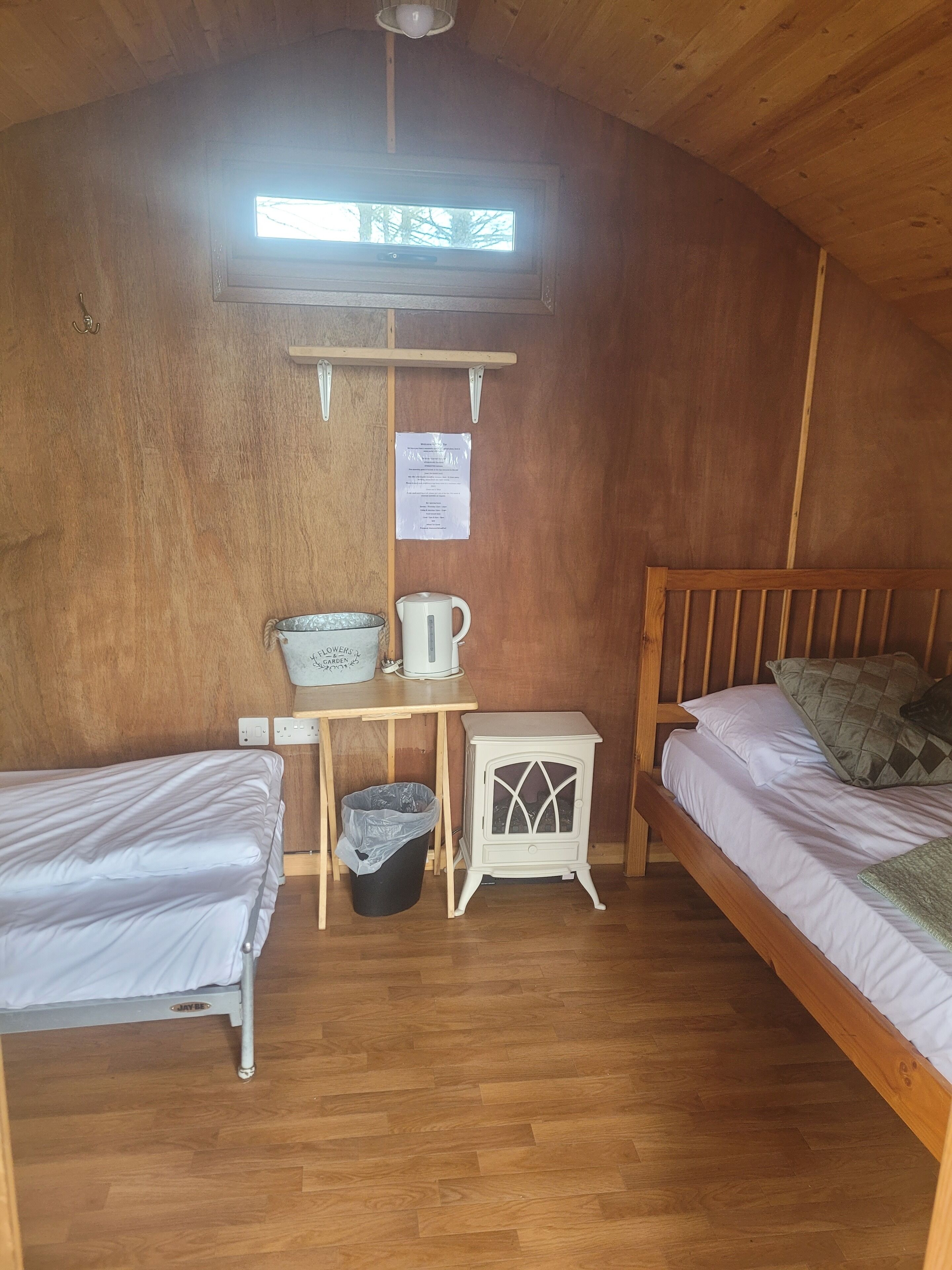 A cozy room with two single beds, a small table with a kettle, a trash bin, and a heater.
