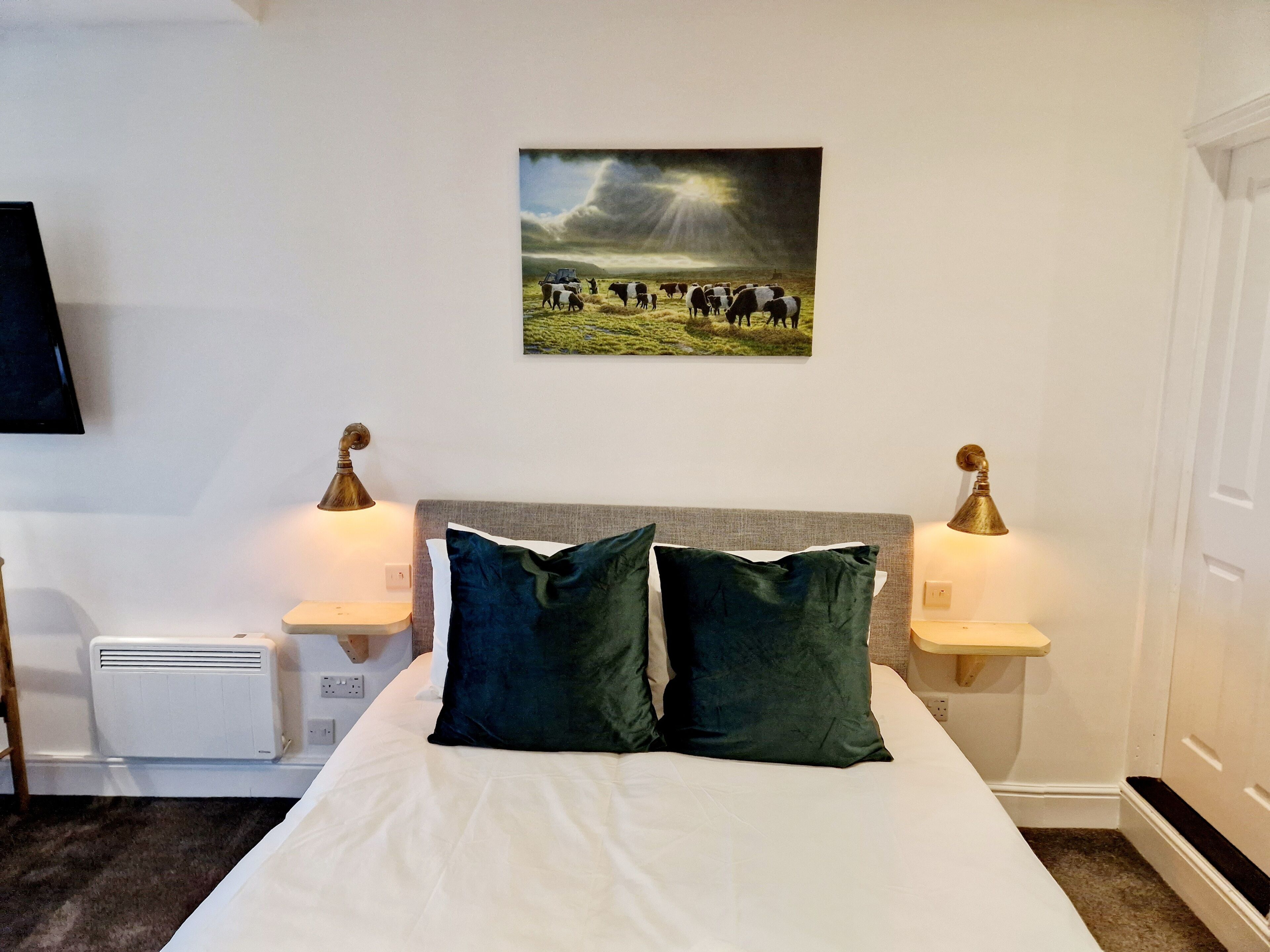 A hotel room with a bed, two bedside lamps, a wall-mounted TV, and a painting of a field with cows.