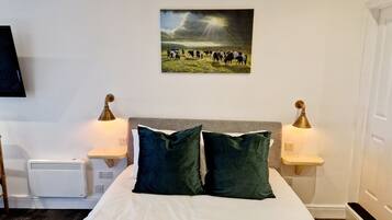 A hotel room with a bed, two bedside lamps, a wall-mounted TV, and a painting of a field with cows.