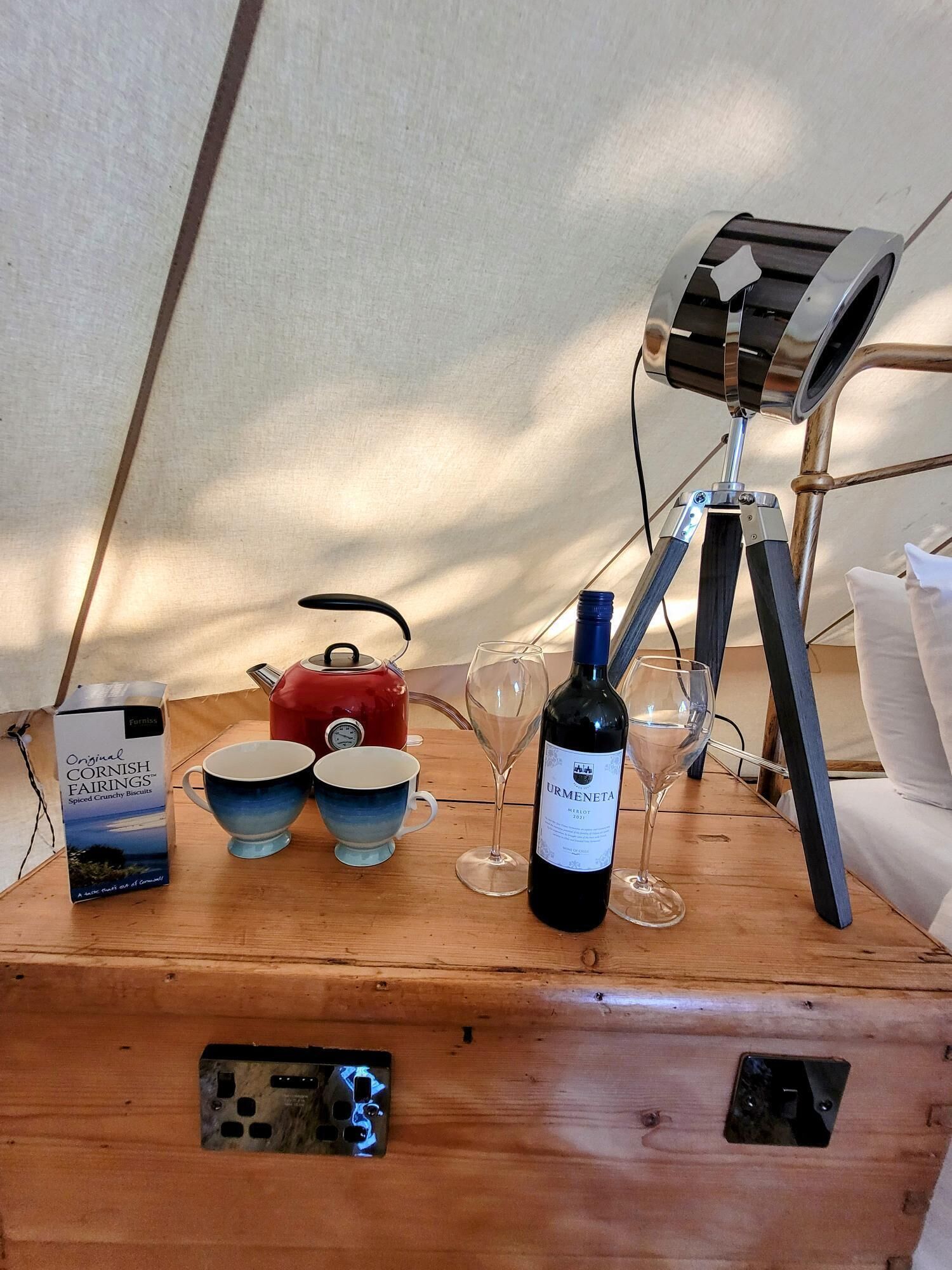 A wooden table with a box of Original Cornish Fairings, a red kettle, two cups, a wine bottle, and a tripod-mounted lamp.