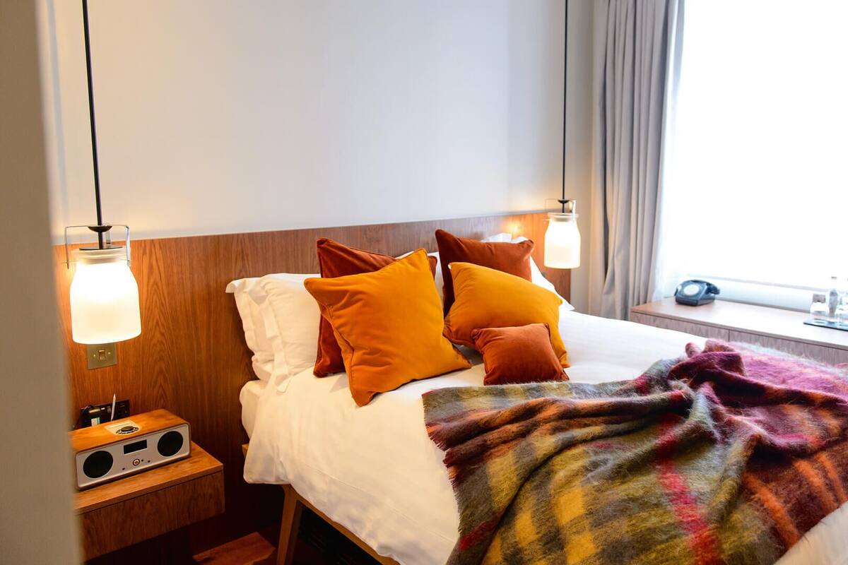 A hotel room with a bed, two bedside lamps, a nightstand with a radio, a window with curtains, and a blanket on the bed.