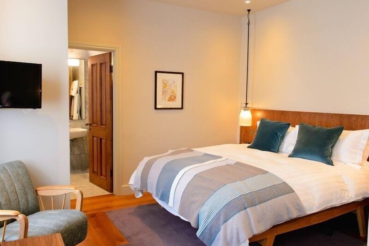 A hotel room with a bed, a chair, a TV, a framed picture, and a door leading to a bathroom.