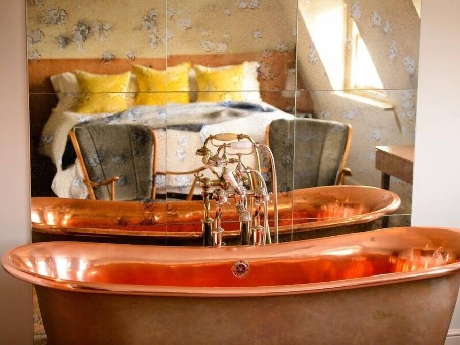 A bathroom with a large copper bathtub, a mirror reflecting a bed with yellow pillows, and a chair.