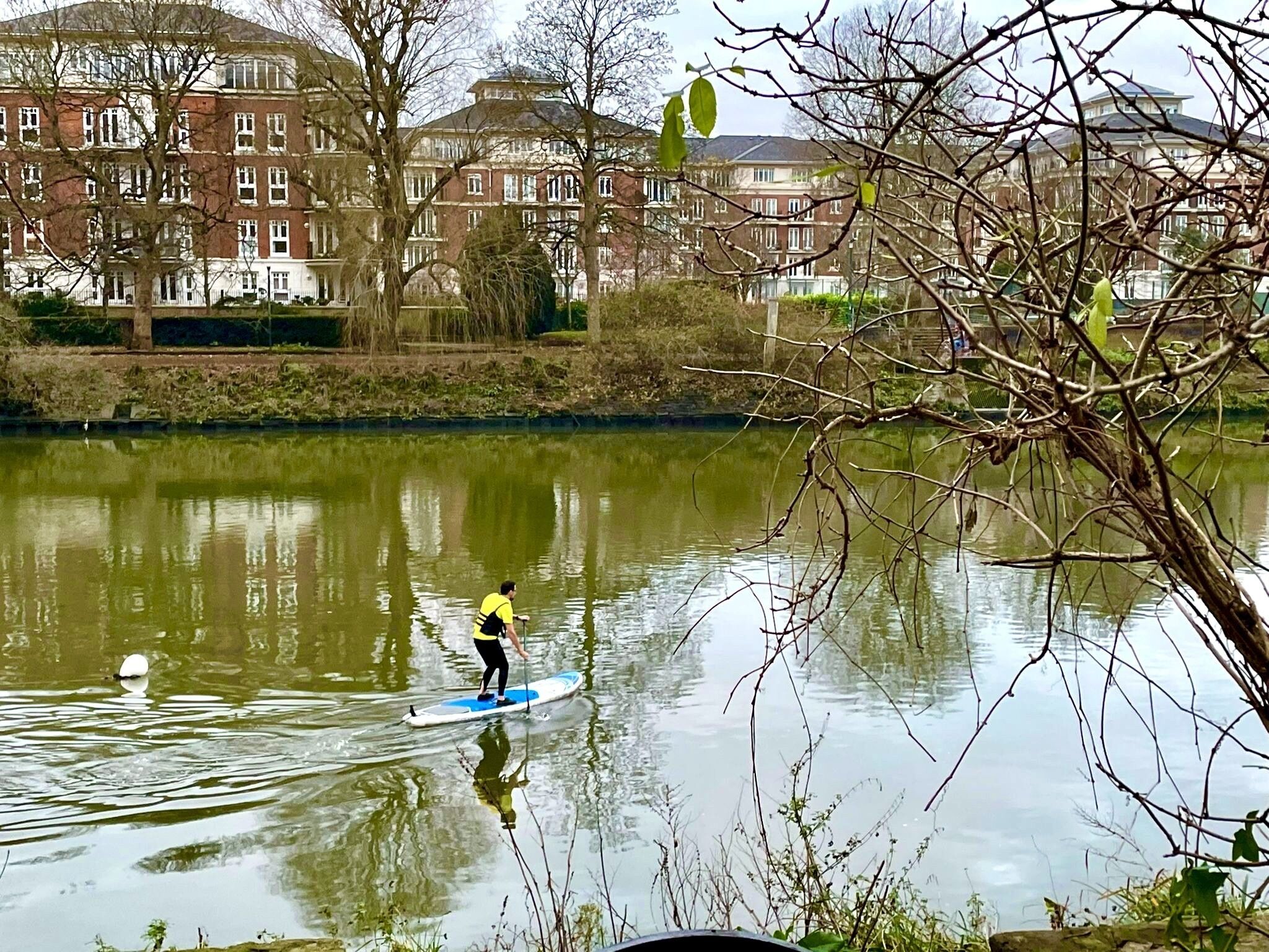 Eine Person steht auf einem Paddleboard auf ruhigem Wasser, im Hintergrund ein Gebäude.