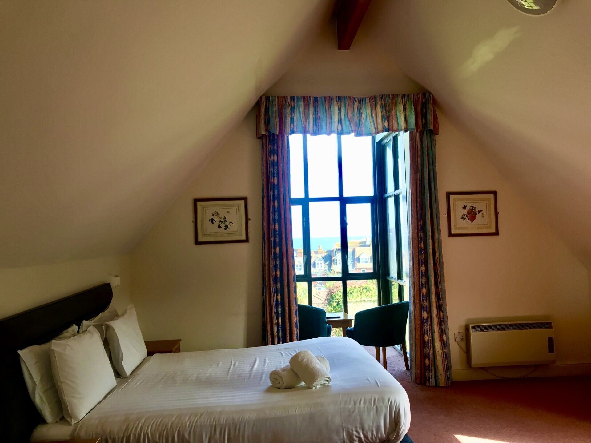 A bedroom with a large bed, a window with curtains, a radiator, and two framed pictures on the walls.