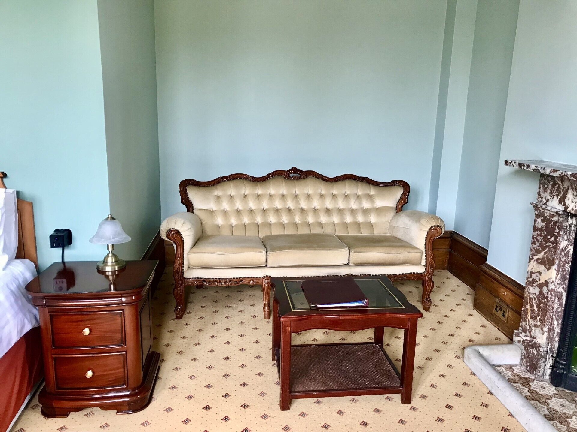 Una habitación con un sofá capitonado, una mesita de noche de madera con cajones, una pequeña mesa y un pilar decorativo.