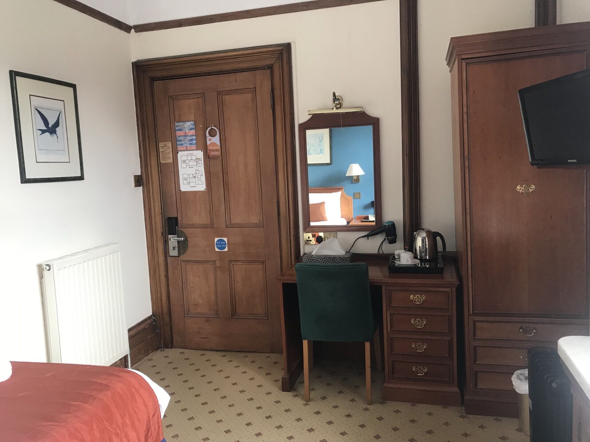 A hotel room with a wooden door, a desk with a chair, a dresser, a radiator, and a television on the wall.