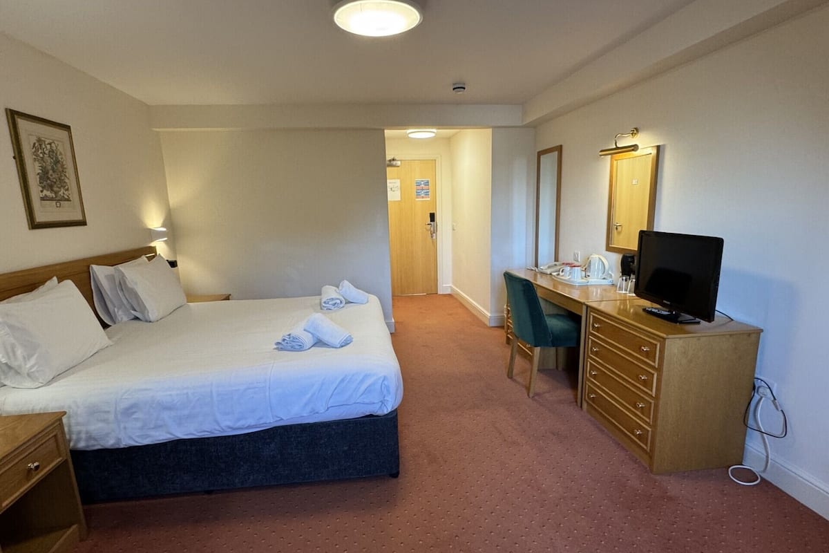 A hotel room with a bed, a desk with a computer and a TV, a dresser, and a door leading to another room.
