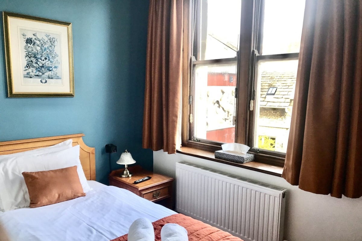 A neatly made bed with white and brown bedding, a framed artwork on the wall, a bedside table with a lamp, and a window with curtains.