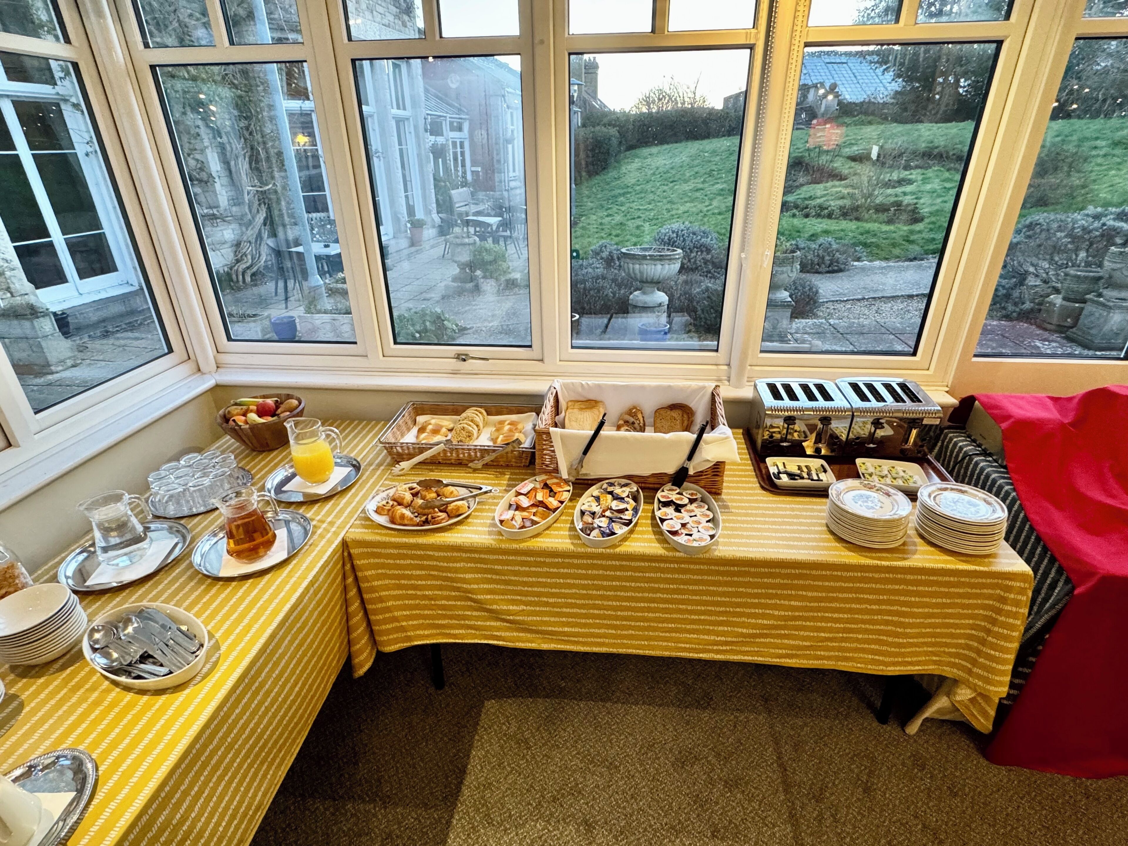 Ein reichhaltiges Frühstücksbuffet mit verschiedenen Speisen und Besteck auf einem Tisch, betrachtet durch große Fenster.