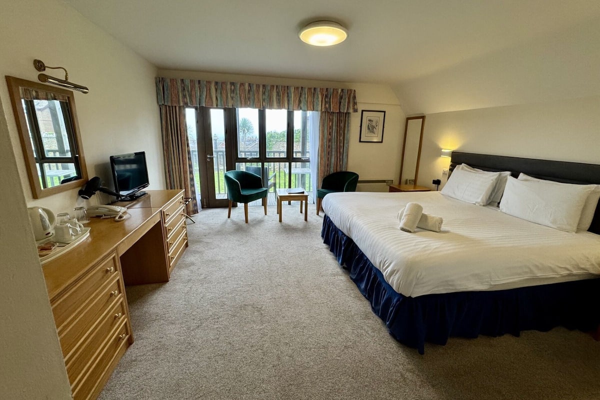 A hotel room with a large bed, a desk with a computer, a chair, and a window with curtains.