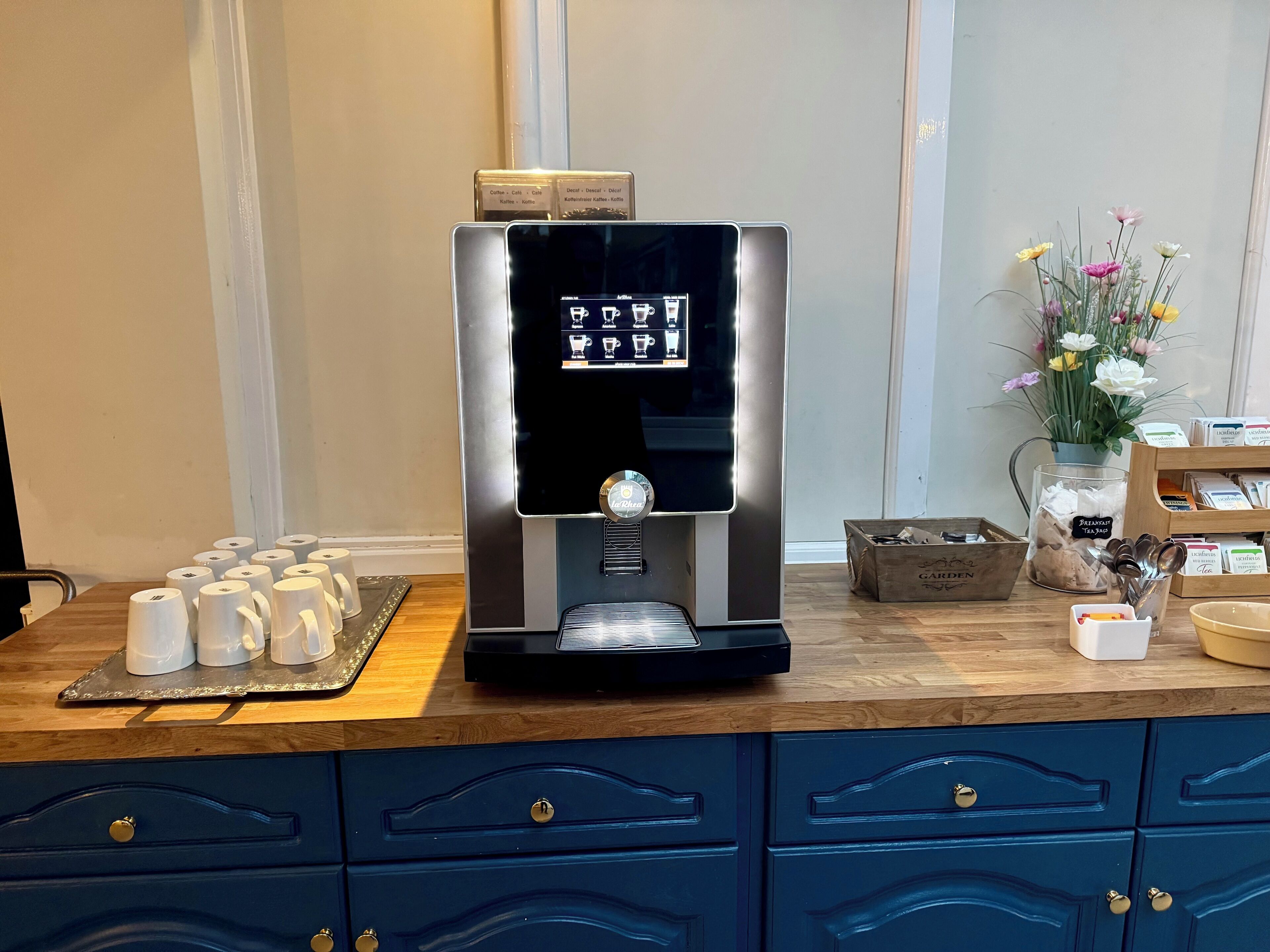 A coffee machine with a digital display, a tray of disposable cups, and a coffee bean container on a wooden counter.