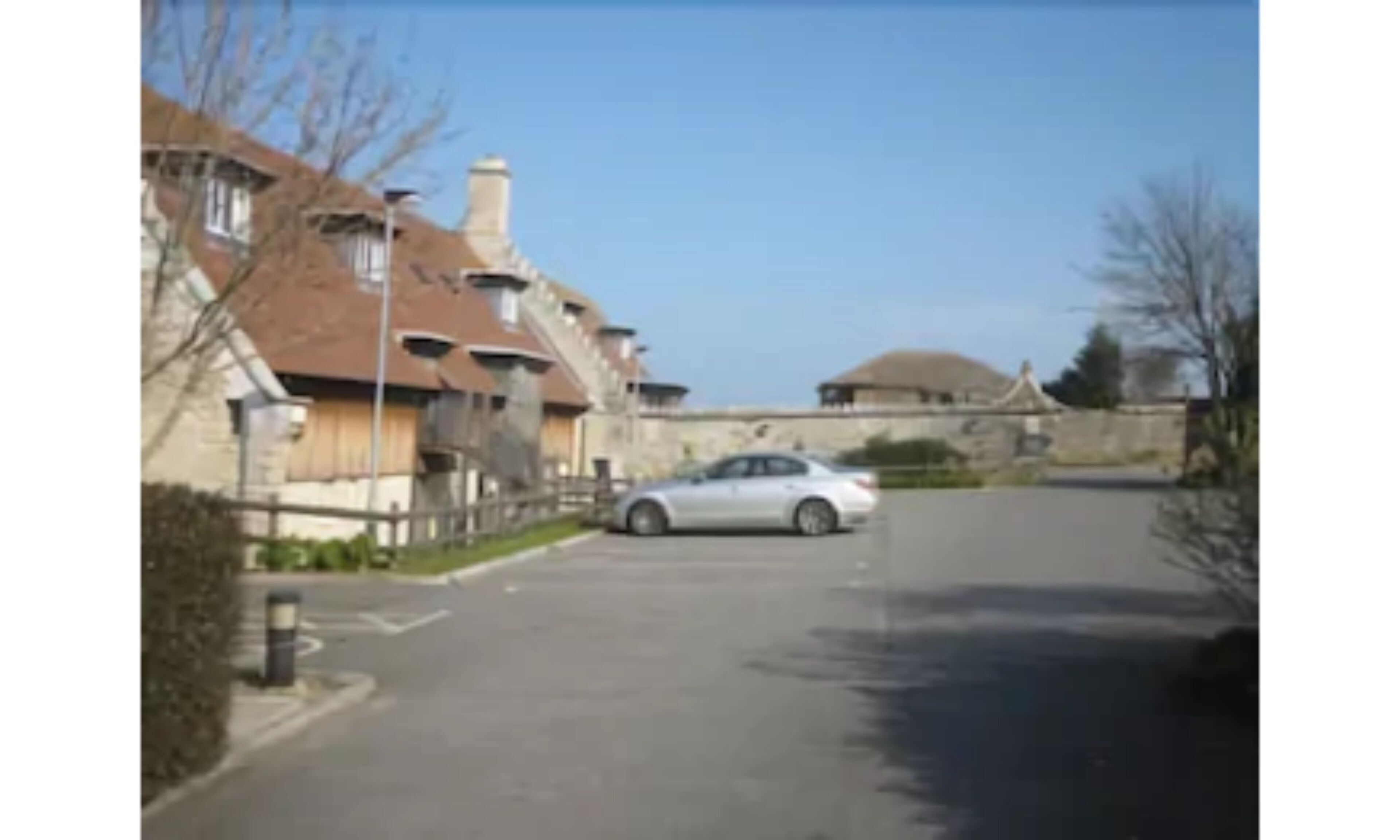 A street view with a white car parked on the side, a stone building with a dome roof, and a clear blue sky.
