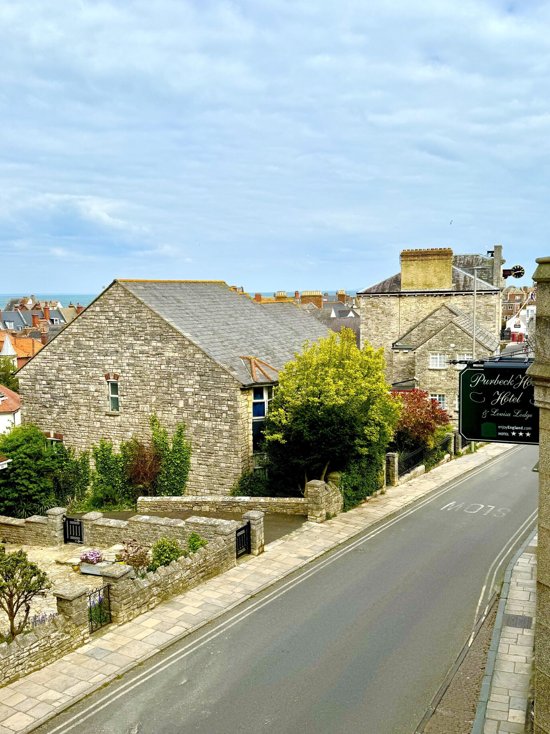 Un bâtiment en pierre avec une enseigne indiquant « Purbeck House Hotel & Louisa Lodge », situé le long d’une rue d’où l’on a une vue dégagée sur la mer.