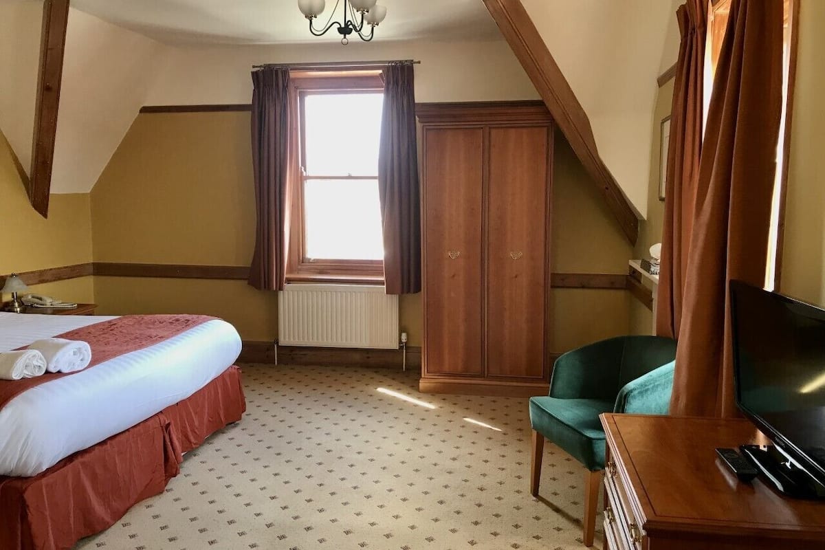 A hotel room with a bed, a chair, a TV, a window with curtains, a radiator, and a chandelier.
