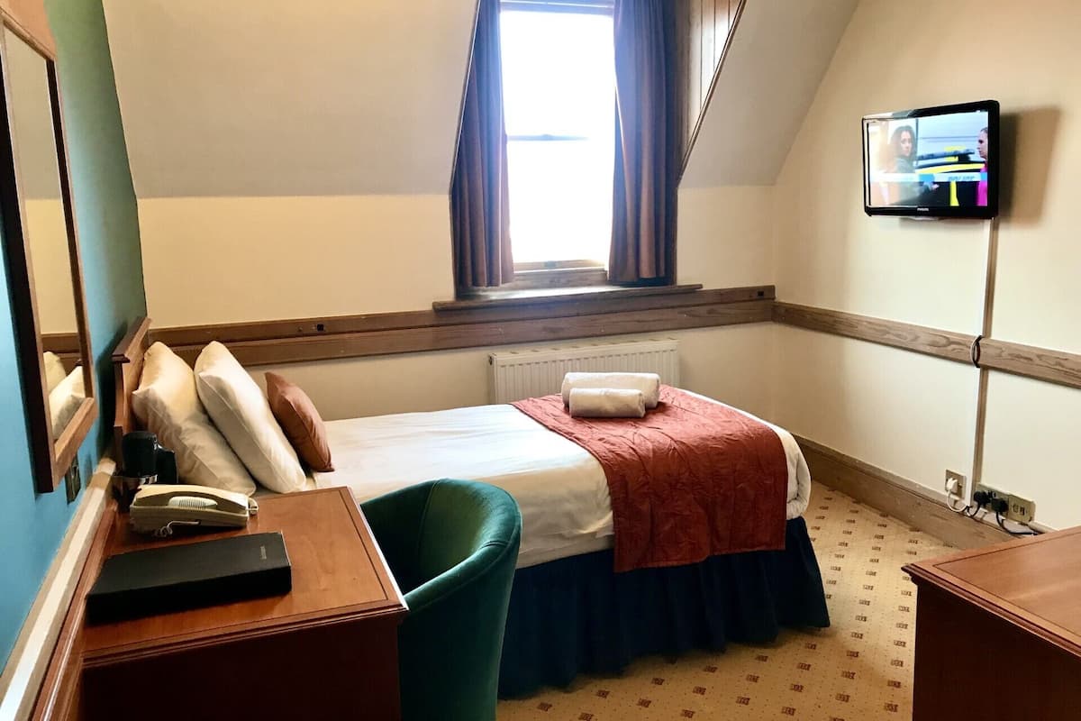 A small, neatly arranged hotel room with a single bed, a desk with a telephone, a chair, and a television mounted on the wall.