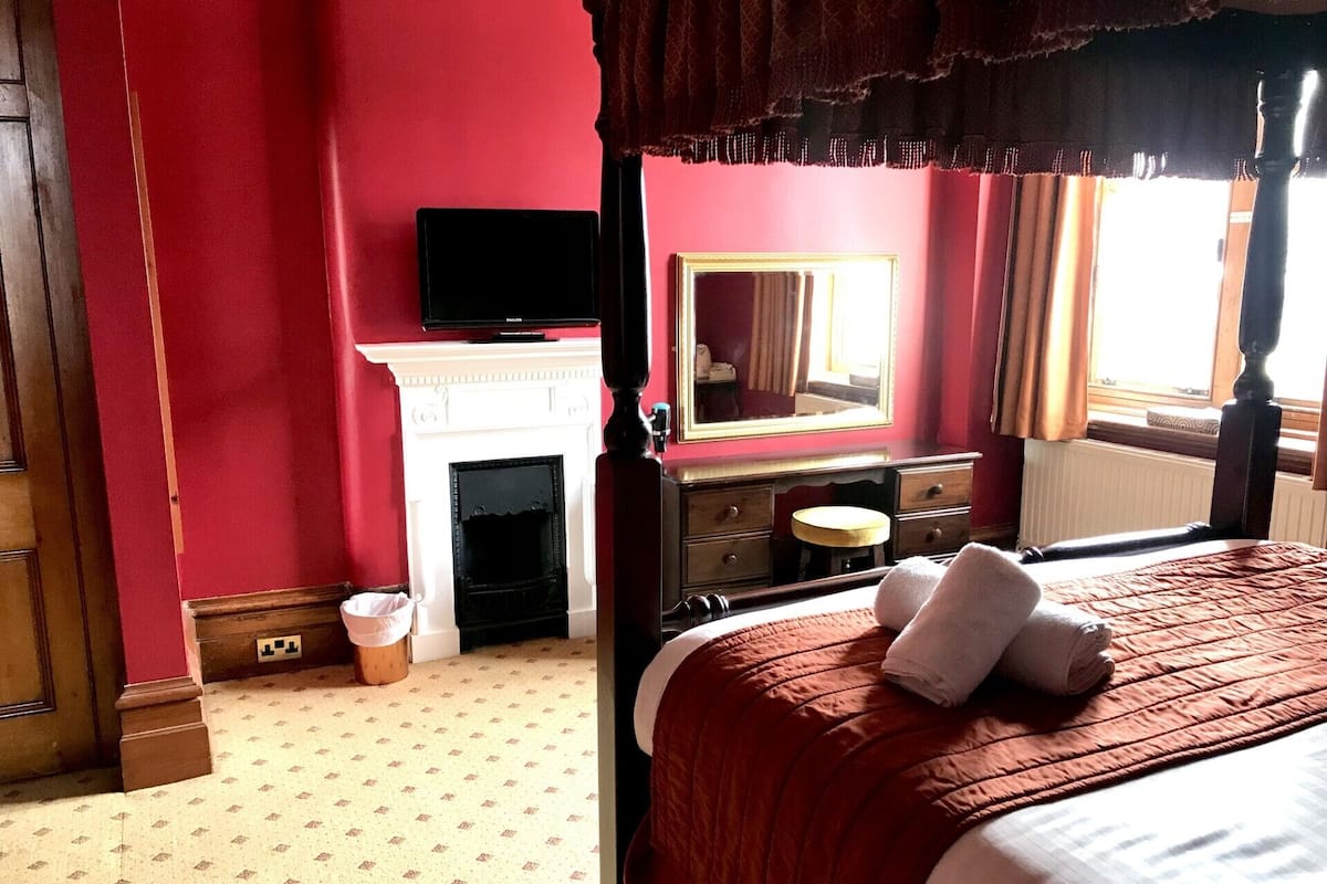 A bedroom with a four-poster bed, a fireplace, a vanity with a mirror, and a television on a stand.