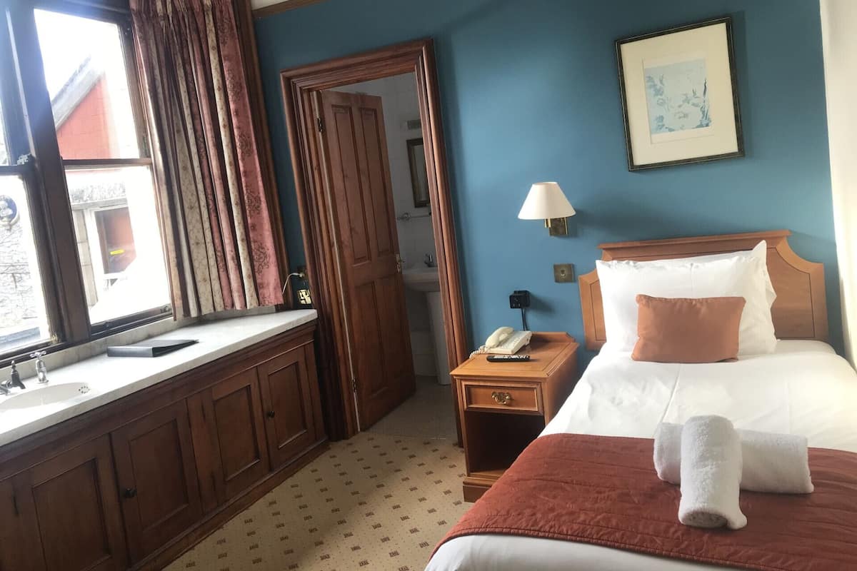 A hotel room with a bed, a nightstand, a bedside lamp, a window with curtains, a bathroom visible through an open door, and a framed picture on the wall.