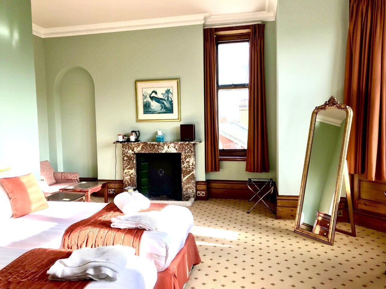 Une chambre d’hôtel avec un lit, une cheminée, un grand miroir et une fenêtre avec des rideaux.