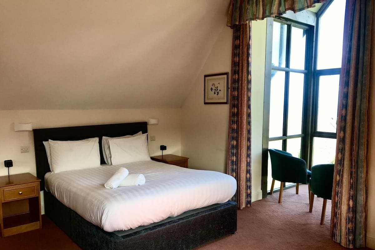 A neatly made bed with white linens and a rolled towel, a bedside table with a lamp, and a chair near a large window with curtains.
