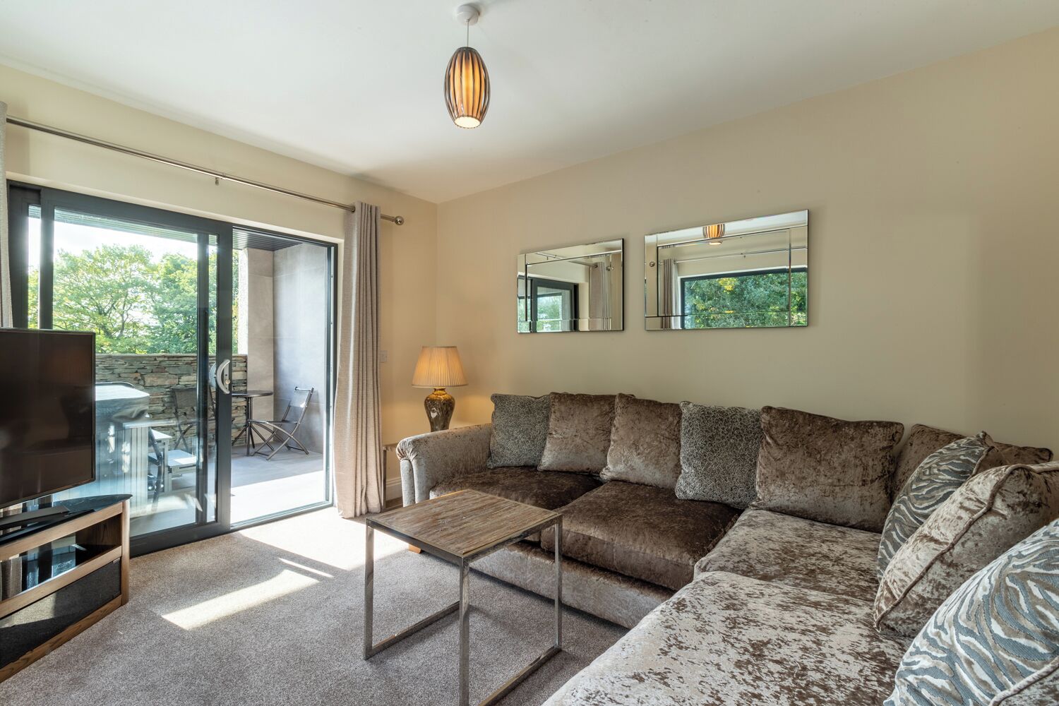 A modern living room with a sectional sofa, a glass coffee table, a flat-screen TV, and a dining area visible through a sliding glass door.