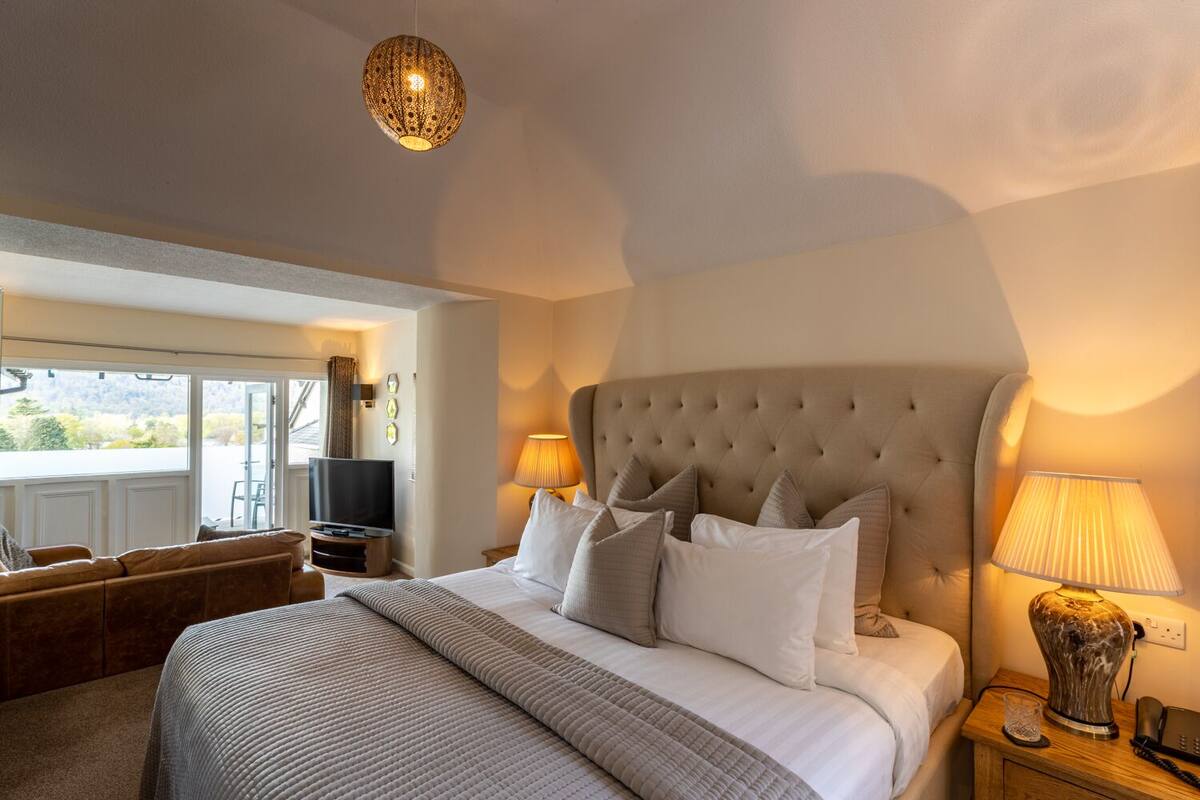 A bedroom with a large bed, two bedside lamps, a television, and a view of the outdoors.