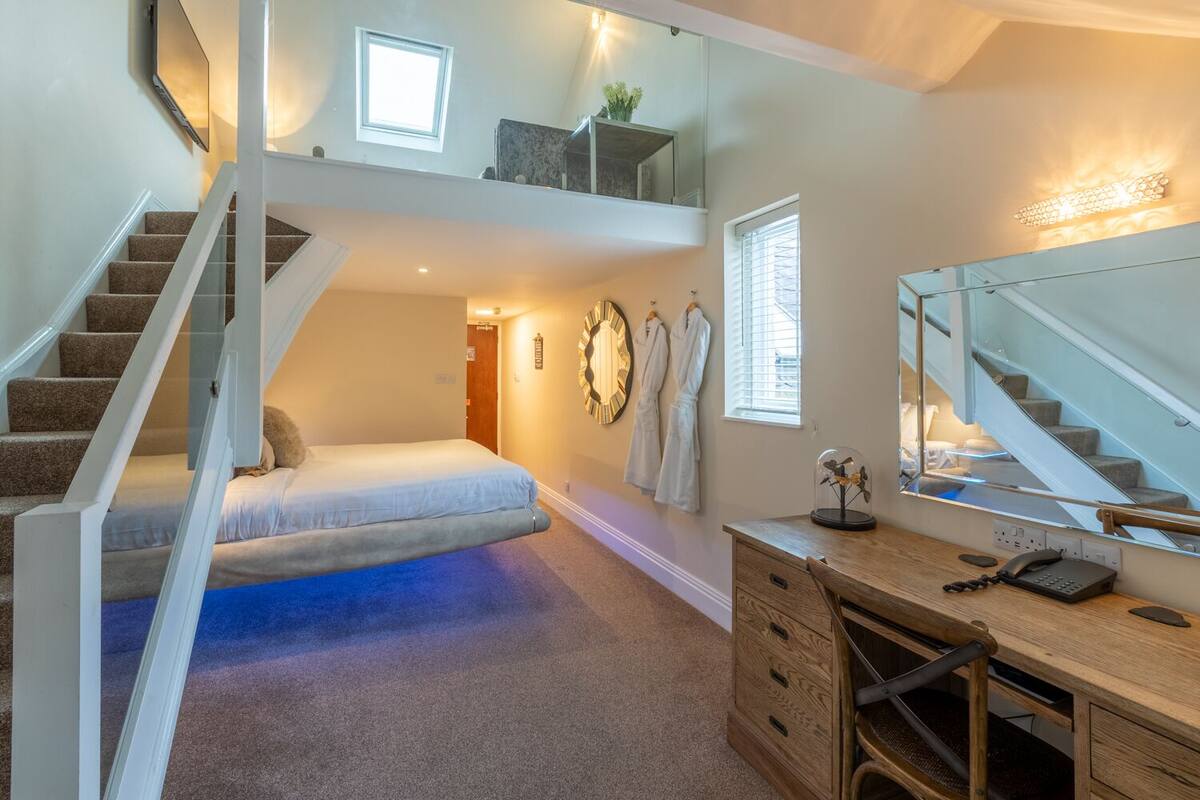 A bedroom with a bed, a desk, a mirror, and a staircase.
