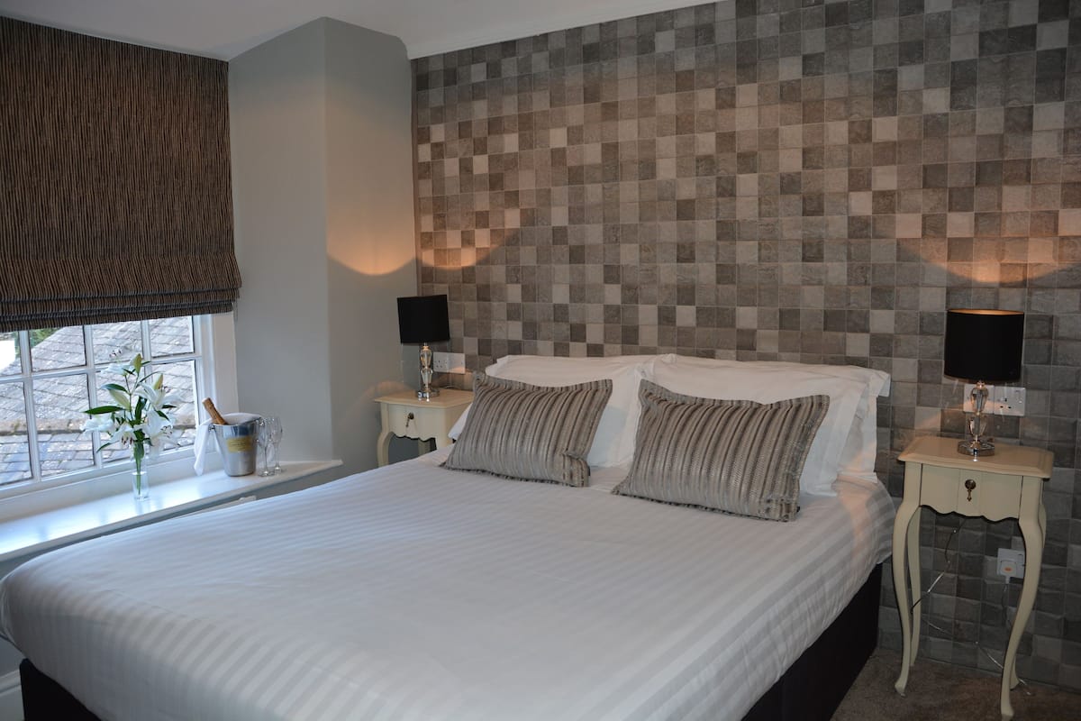 A neatly made bed with pillows, a bedside table with a lamp, a window with a view, and a wall with a checkered pattern.