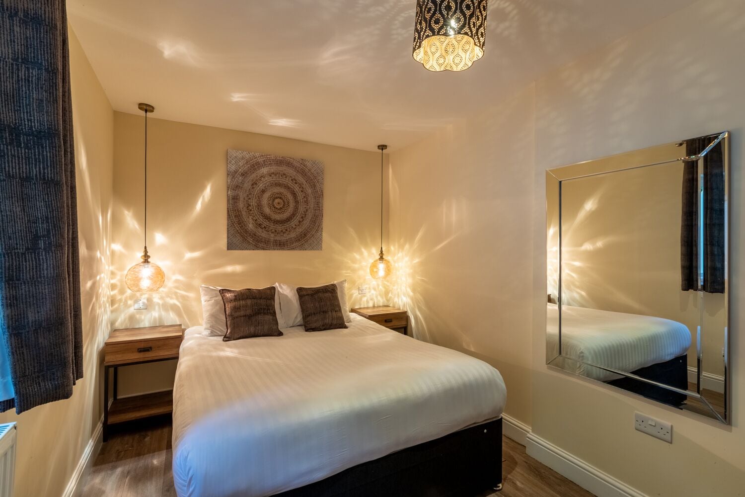 A bedroom with a bed, two bedside tables, a mirror, a wall tapestry, and pendant lights.