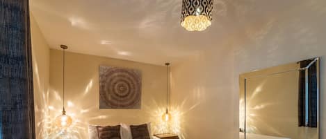 Une chambre à coucher comprenant un lit, deux tables de chevet, un miroir, une tapisserie murale et des lampes suspendues.
