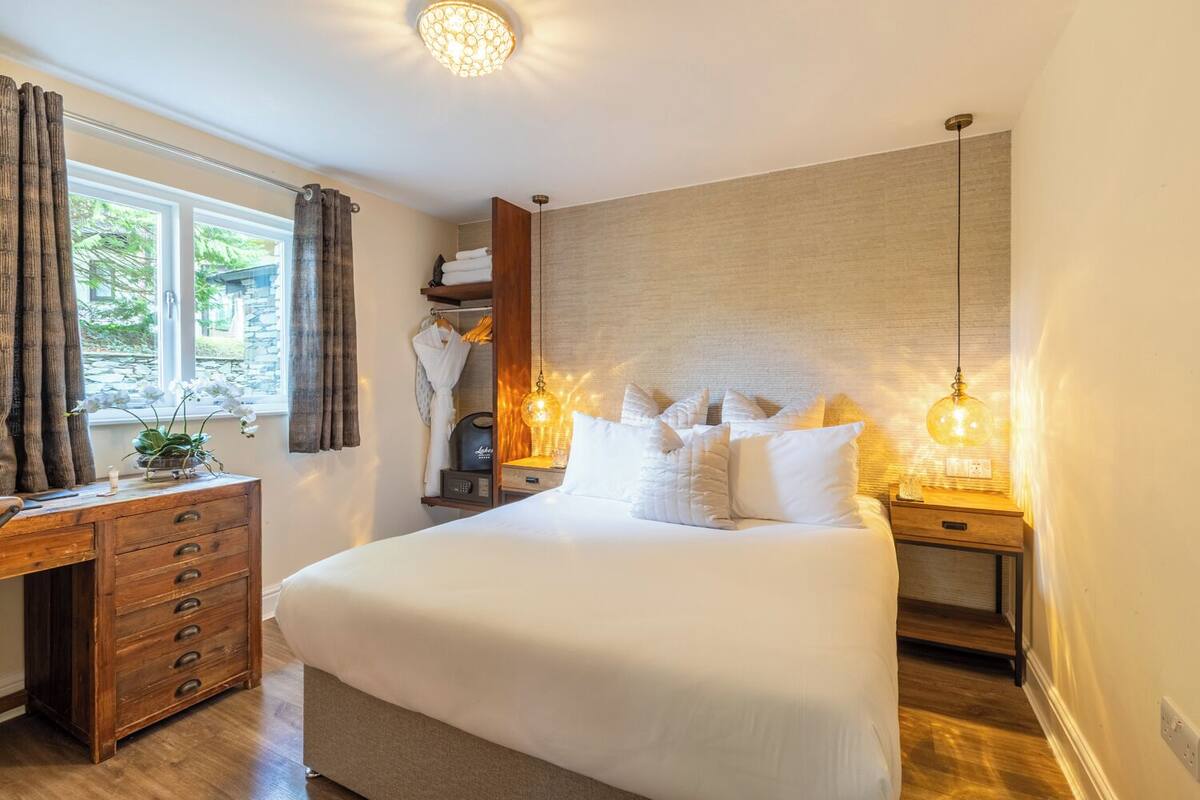A spacious bedroom with a large bed, wooden dresser, and a desk. There is a window with curtains, a bedside table with a lamp, and a wall-mounted shelf with towels.