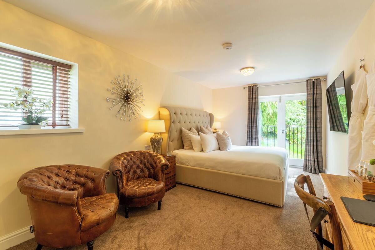 A bedroom with a large bed, two armchairs, a bedside table with a lamp, a decorative wall piece, and a window with blinds.
