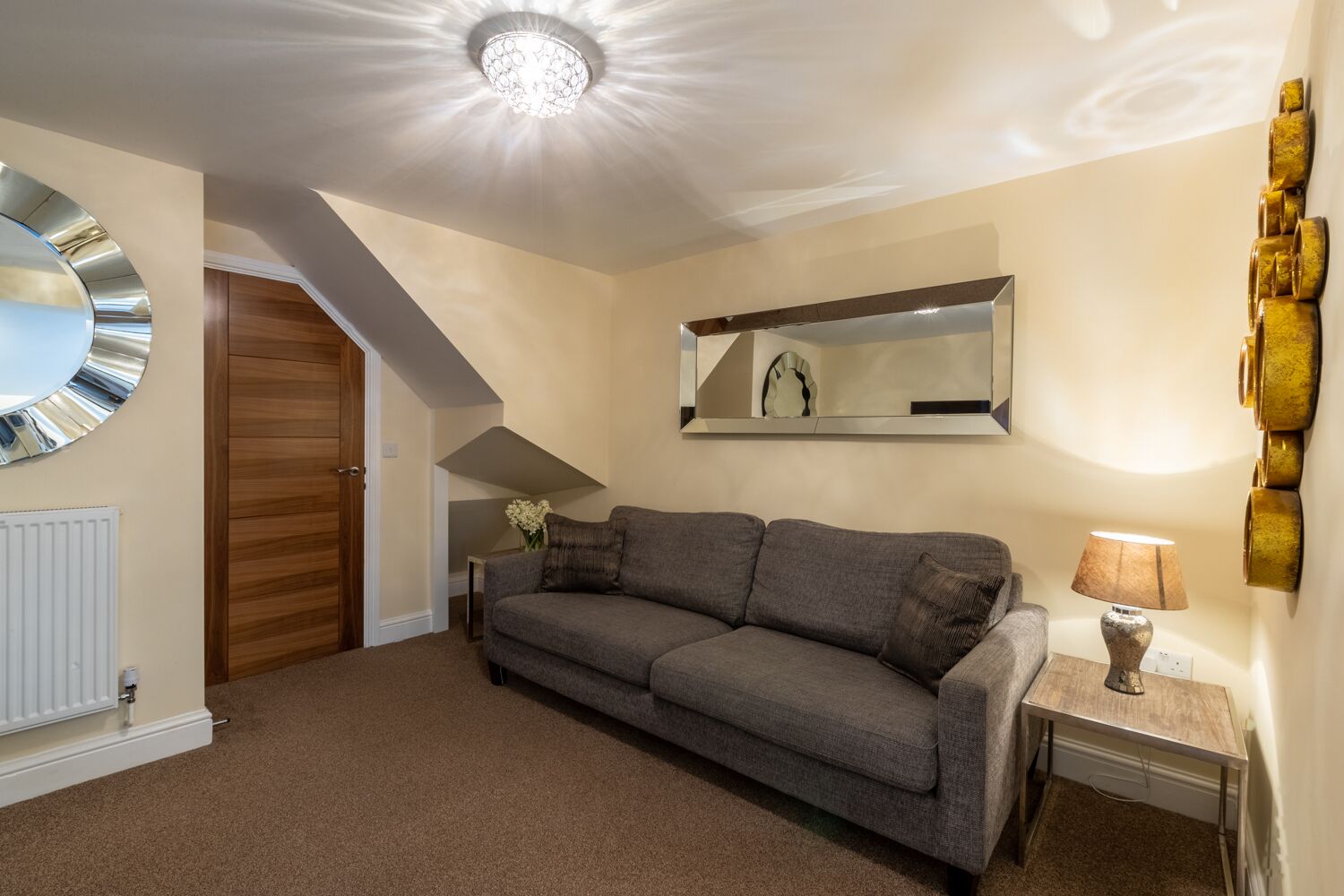 A living room with a grey sofa, a wooden door, a white radiator, a mirror, and a lamp on a side table.