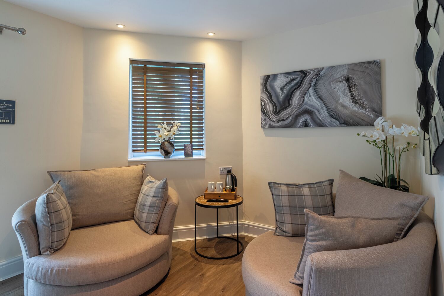 A modern living room with two beige armchairs, a small round coffee table, and a window with blinds.