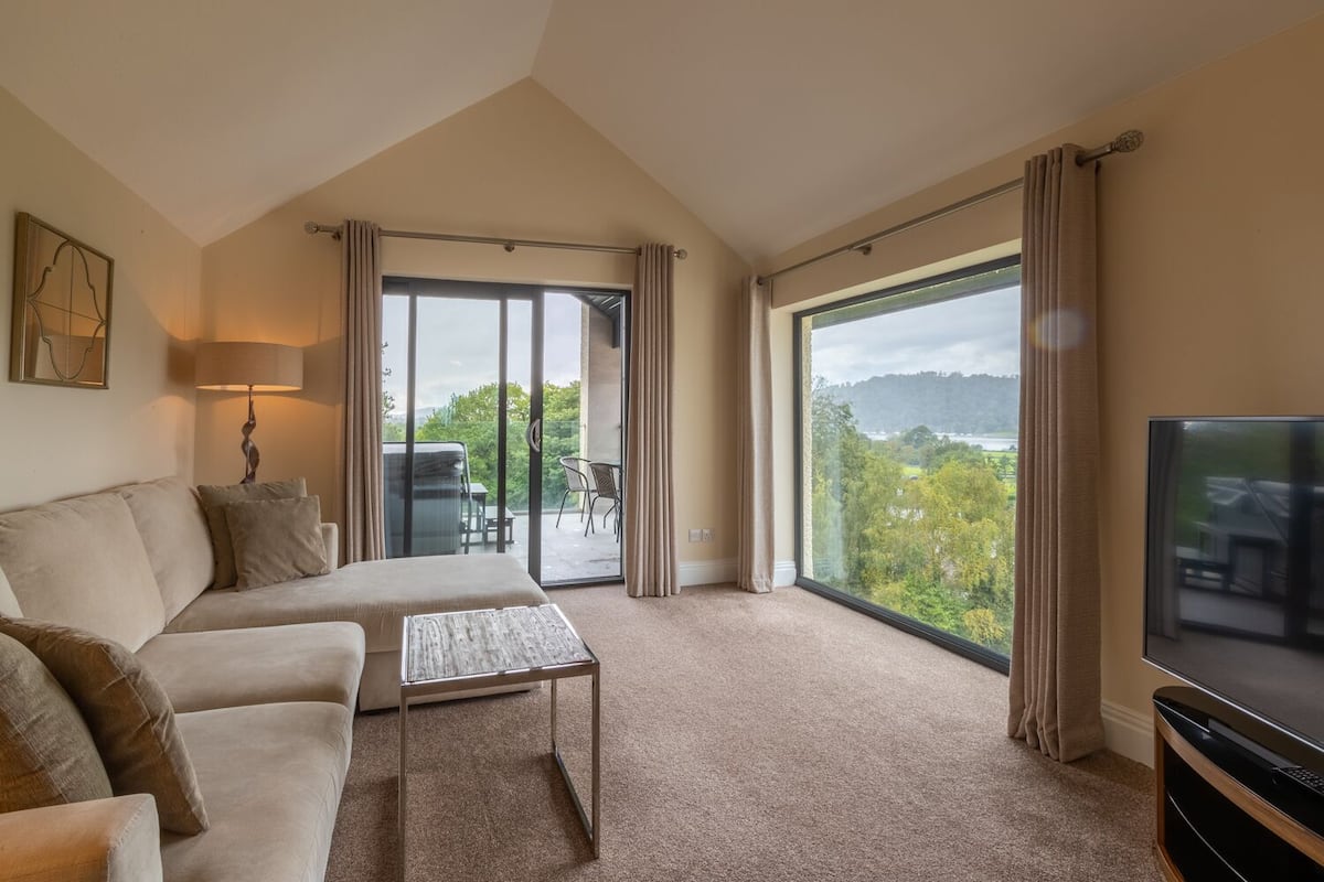 A spacious living room with a sofa, a coffee table, and a TV. There is a large window offering a view of greenery and a body of water.