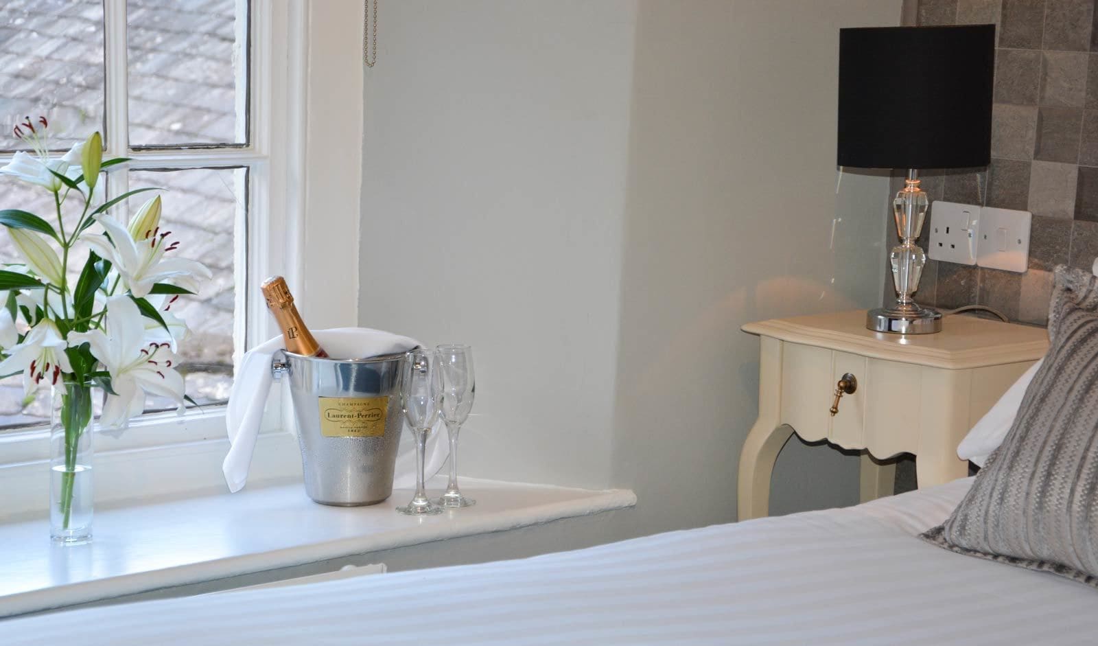 Habitación de hotel con cama, mesita de noche con lámpara, ventana con vistas, un jarrón con flores y un cubo de champán con copas.