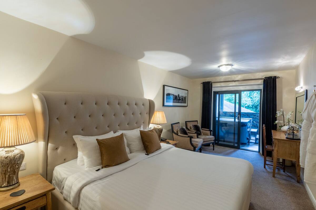 A hotel room with a large bed, two bedside lamps, a wooden nightstand, a chair, a desk, and a view of the outdoors through a glass door.