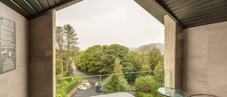 A balcony with a glass table and chairs, a view of a road and trees, and a sign on the wall.