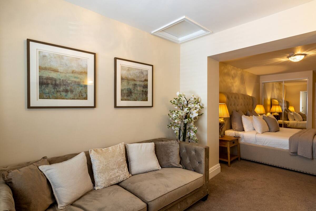 A living room with a beige sofa, two framed abstract paintings, a vase of white flowers, and a bedroom with a bed, lamps, and a mirror.