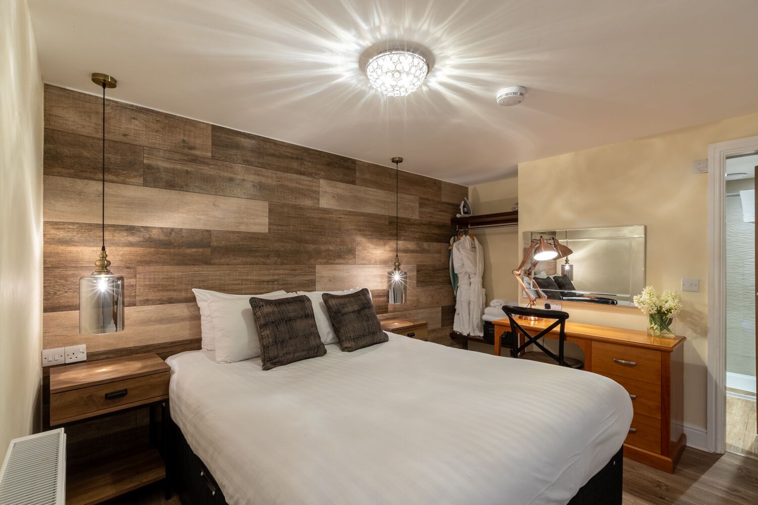 Une chambre d’hôtel moderne avec une tête de lit en bois, un grand lit avec du linge de lit blanc, un bureau avec un miroir et un porte-peignoir.