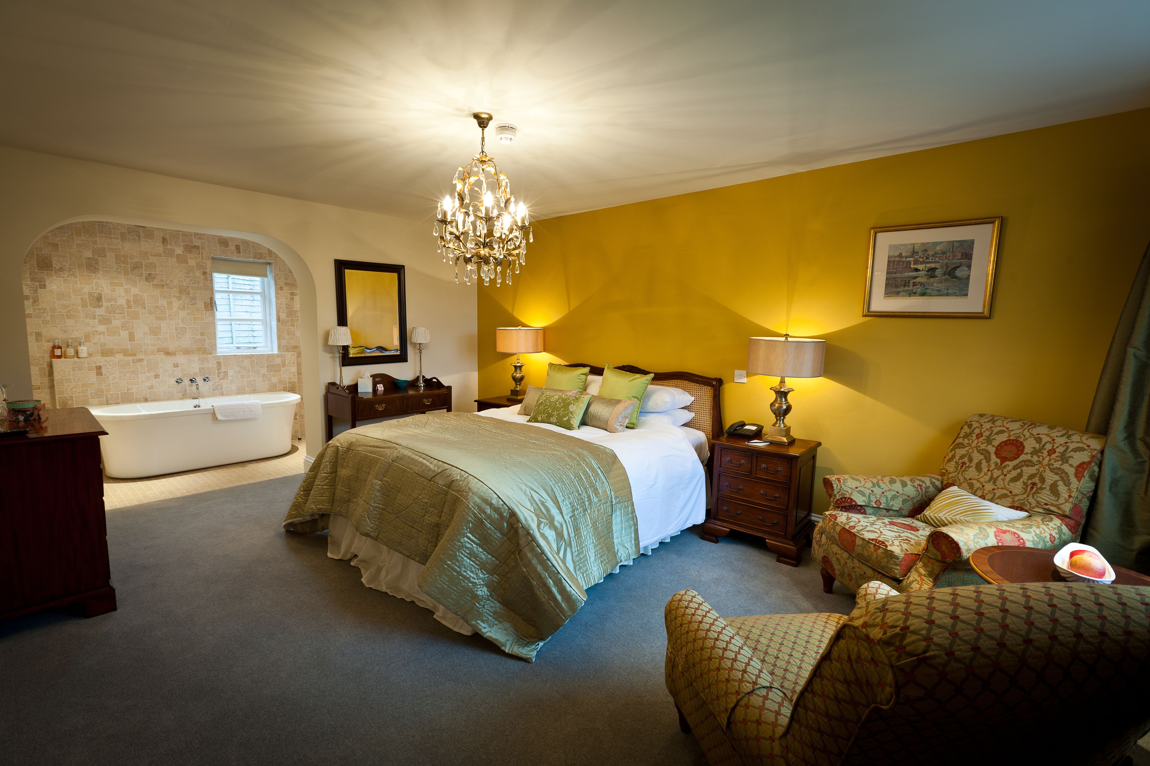 A bedroom with a large bed, a sitting area with a chair, a small table, a nightstand, a lamp, a mirror, a picture on the wall, and a bathtub.