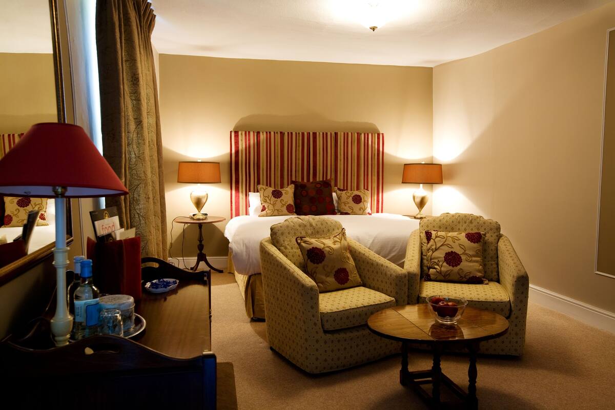 Ein Hotelzimmer mit einem Bett, zwei Sesseln, einem kleinen Tisch, einer Lampe, einem Spiegel und einem Nachttisch mit Lampe.