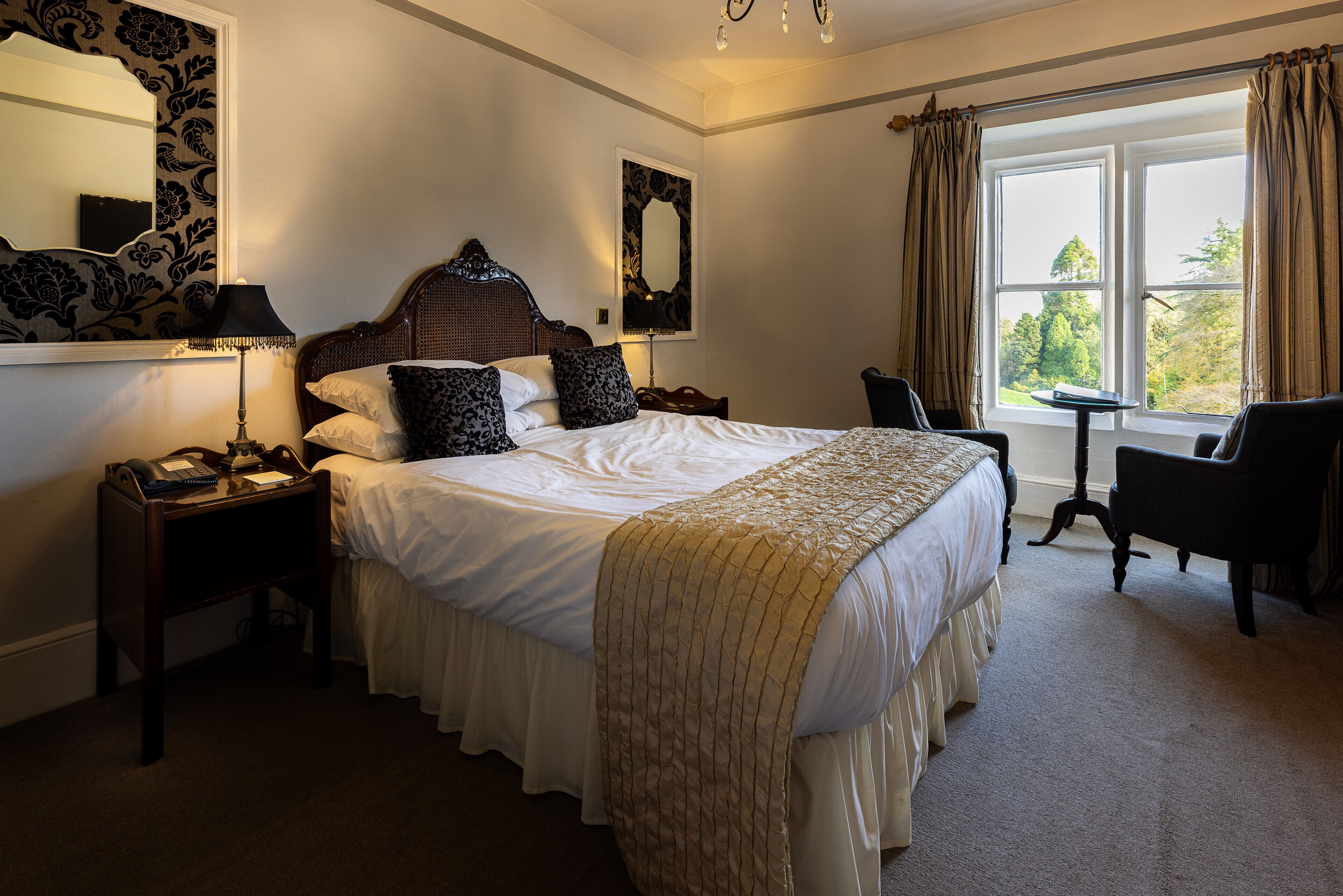 A hotel room with a large bed, a nightstand, a desk, a chair, a window with curtains, and a mirror on the wall.