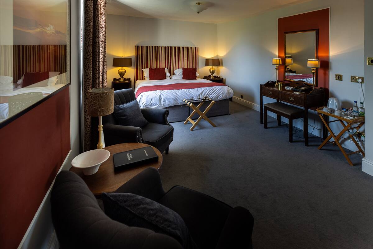 Ein Hotelzimmer mit einem großen Bett, zwei Nachttischen mit Lampen, einem Schreibtisch mit Stuhl, einem kleinen Tisch ebenfalls mit Lampe und einem Spiegel an der Wand.