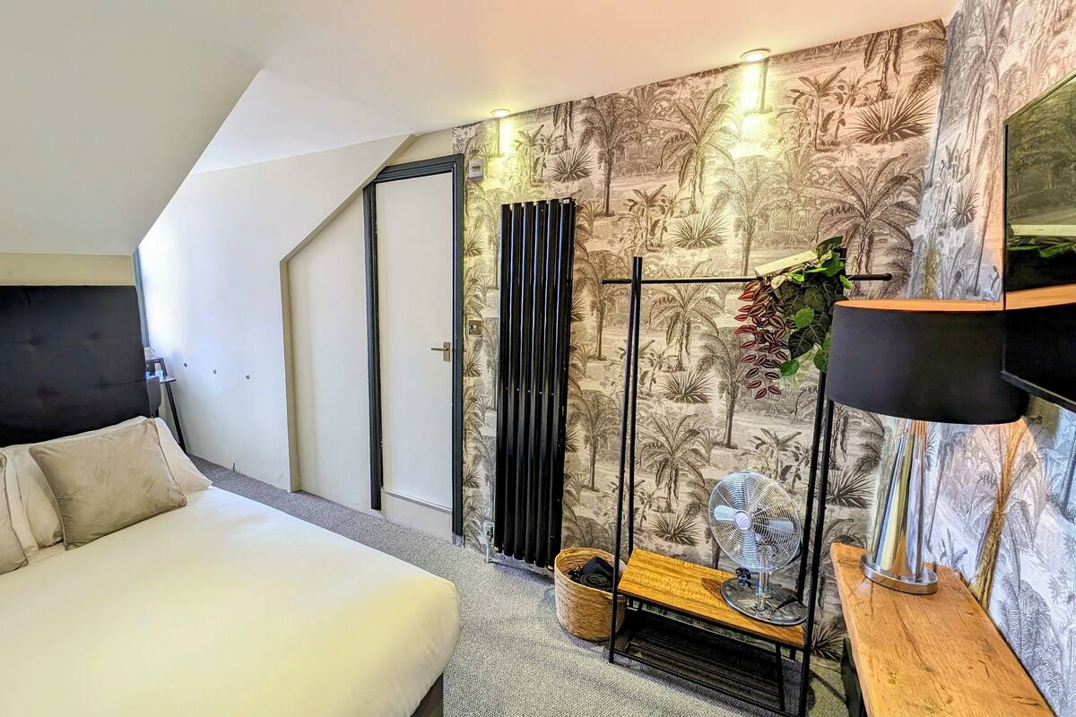 A modern bedroom with a white bed, a black headboard, a wooden side table, a floor lamp, and a wall-mounted radiator.