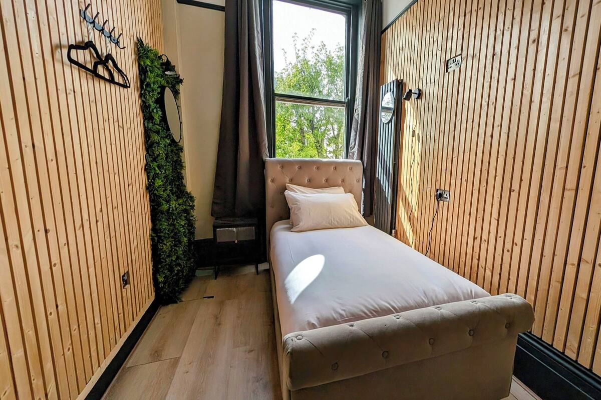 A narrow room with wooden paneling, a bed with white bedding, a mirror, and a small potted plant.
