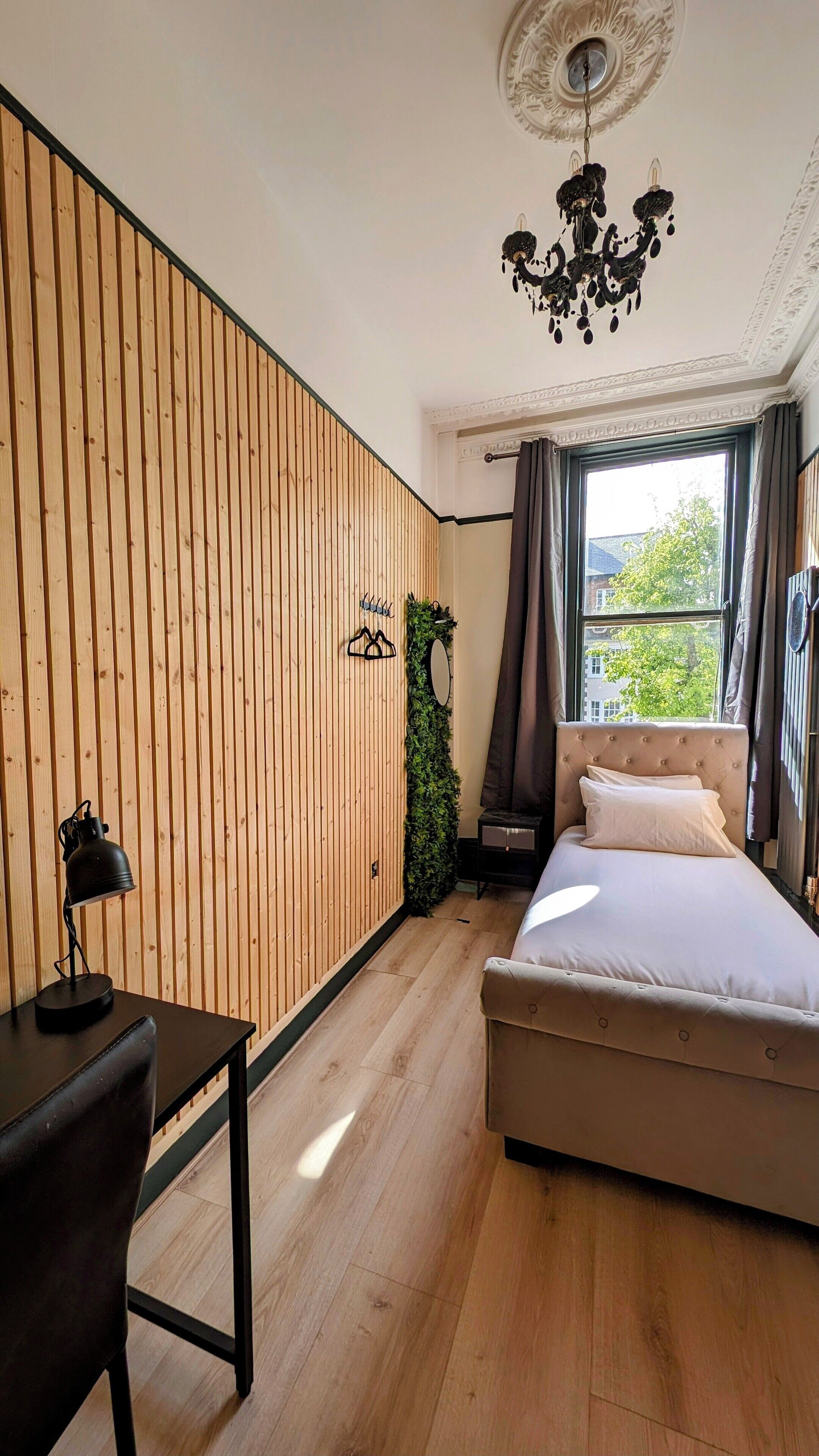 A bedroom with a wooden panel wall, a bed with a tufted headboard, a desk with a lamp, a hanging plant, and a window with curtains.