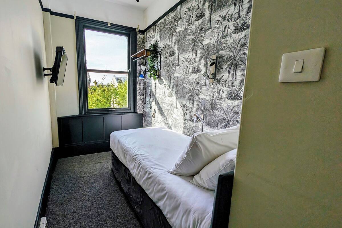 A small bedroom with a bed, a window, and a wall with a tropical wallpaper.