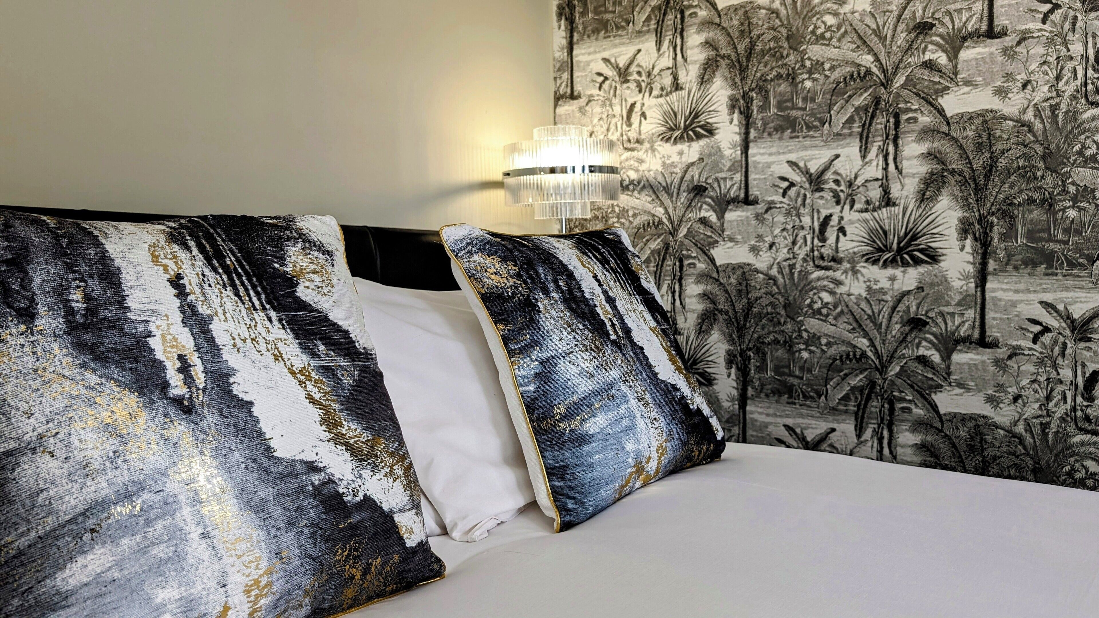 Et hotelværelse med en seng, to puder med et sort-hvidt abstrakt design, og et vægmaleri af et tropisk landskab.